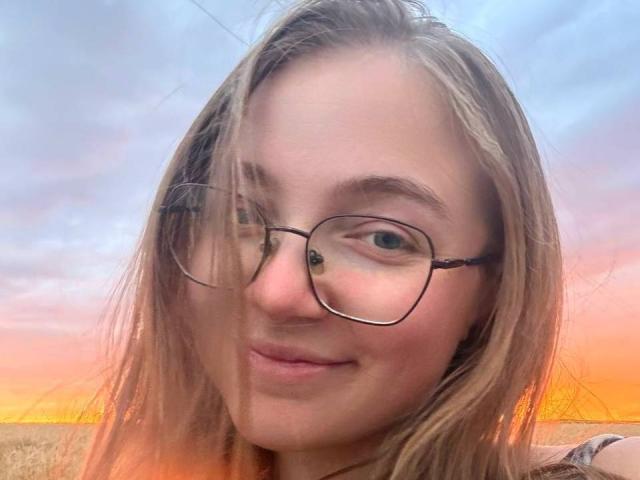 ArrinaWair has light brown hair, glasses, and a subtle smile, with a slight wind in her face, showing a natural and cute look. Her tits are small, not visible.