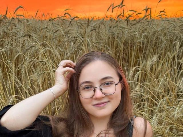 ArrinaWair has brown hair, glasses, and a slight smile, with her hand in her hair, standing in a wheat field at sunset. She's wearing a black top.