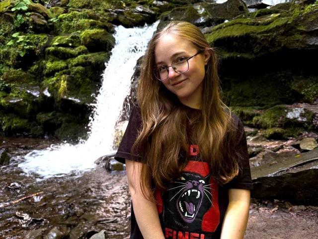 ArrinaWair, with long brown hair and glasses, wears a black T-shirt with a red and black panther, standing in front of a small waterfall in a mossy forest.