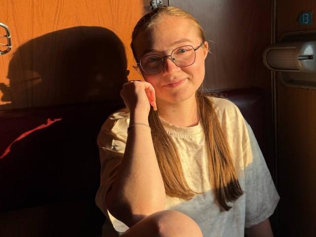 ArrinaWair with blonde pigtails, glasses, and a smile, wearing a white t-shirt, sitting in sunlight, touching her chin, soft shadows, wooden wall, relaxed, cozy atmosphere.