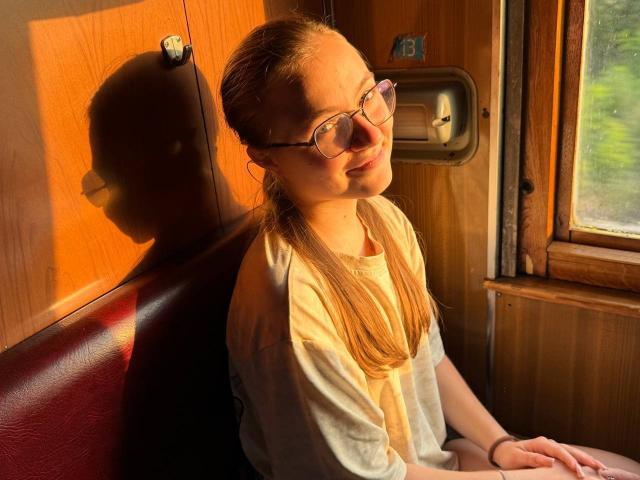ArrinaWair with glasses, blonde hair in a ponytail, white shirt, sitting in sunlight, shadow on wall, wooden window, relaxed, hands on lap, warm orange light.