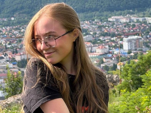 ArrinaWair with long blonde hair, glasses, and a black shirt, smiling at the camera, her small tits visible under her shirt. She's standing in a green, hilly area.