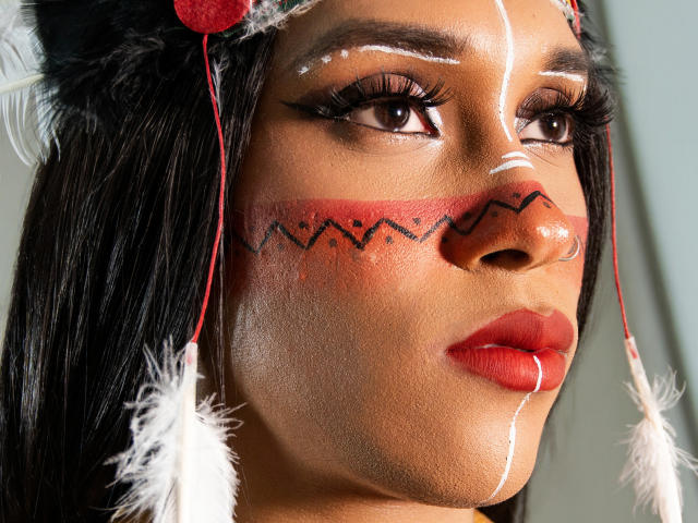 PaulaHills has dark skin, red lipstick, black hair, and white and red face paint with zigzag patterns, wearing a feathered headpiece. Her eyes are large and she has long lashes.