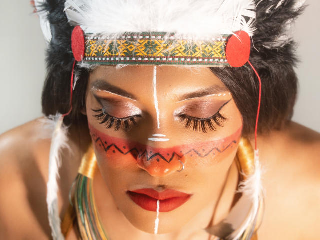PaulaHills with painted face, white feather headband, red nose ring, black feather ears, red lipstick, white and red face paint, and long eyelashes, looking down with eyes closed.