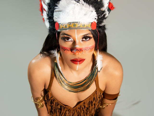 PaulaHills with tan skin, dark eyes, red lipstick, white and black feathered headdress, red face paint, gold necklace, brown fringed top, and gold arm bands, looking up.