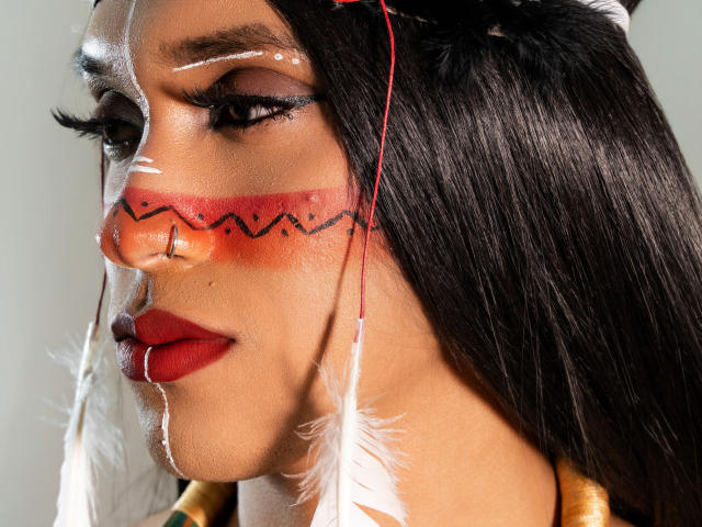 PaulaHills' face with red zigzag paint on nose, white feathers, black hair, red lipstick, and white facial dots. She has a nose ring and long eyelashes.