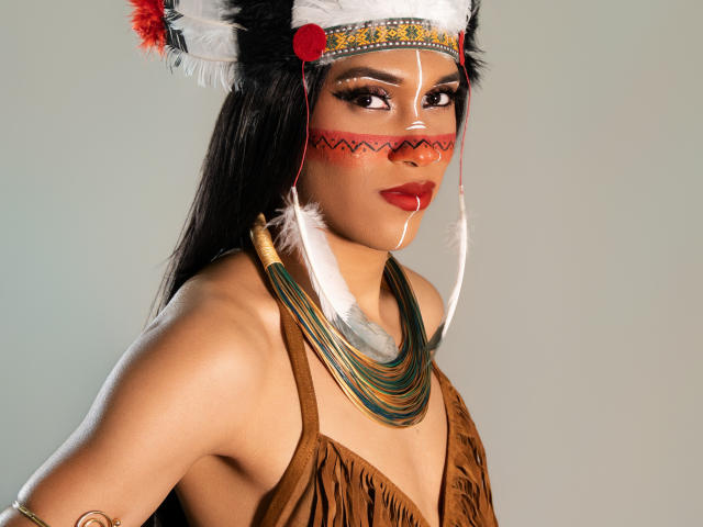 PaulaHills with dark hair, wearing a Native American-style headdress, brown fringed top, red lipstick, white face paint, and multicolored necklace, showing her tits and ass.