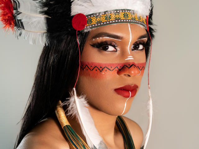 PaulaHills with dark hair, wearing a Native American-style feathered hat, red face paint, and white facial markings, has red lipstick and a nose ring, looking to the side.