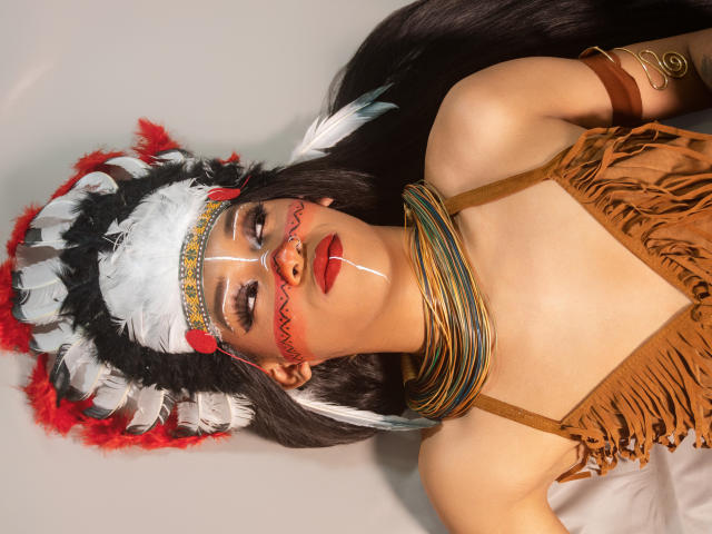 PaulaHills with long black hair, wearing a Native American-style headdress, red and white feathers, brown fringed top, and colorful neck beads, has red makeup on her face and lips.