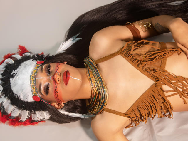 PaulaHills with long black hair, wearing a brown fringed top, feathered headdress, face paint, red lipstick, striped necklaces, and arm band, lying down, looking at camera.