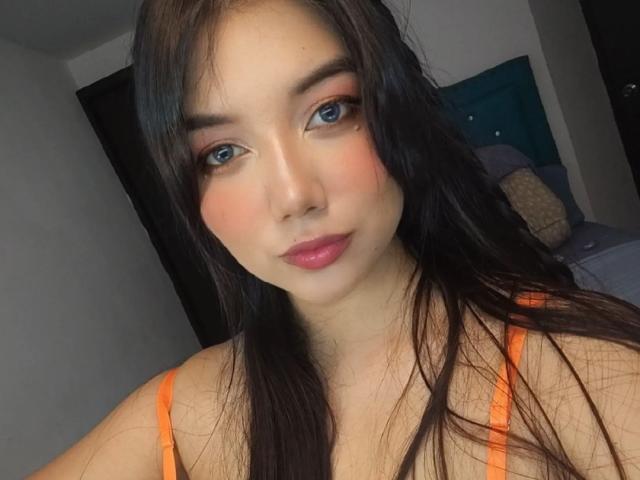 SquirtinGamy has long black hair, blue eyes, pink blush, and pink lipstick. She's wearing an orange top, showing her tits, and looking directly at the camera with a slight smile.