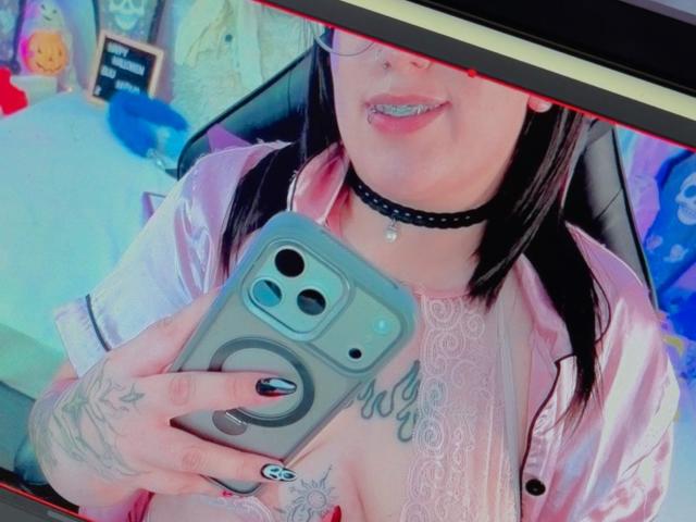 NatalieHarris with black choker, glasses, and grey lipstick, taking a selfie in a mirror, showing tattoos, pink lace bra, and pink satin jacket, holding a grey phone.