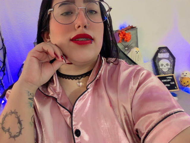Natalie Harris con gafas, lápiz labial rojo, camisa de satén rosa, collar negro, piercing en la nariz, brazo tatuado, uñas rojas, mano cerca del rostro, cabello oscuro, piel pálida, boca ligeramente abierta..