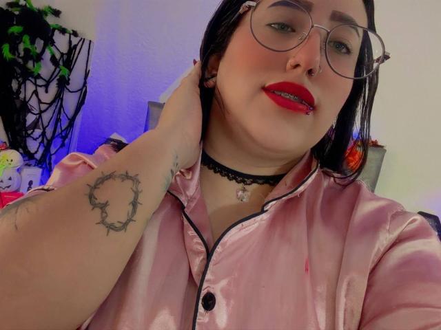 Natalie Harris, con cabello negro, gafas, lápiz labial rojo, piercing en la nariz, collar, camisa de satén rosa, tatuaje en el antebrazo derecho, mano tocándose la cabeza, mirando a la cámara, linda, sexy..