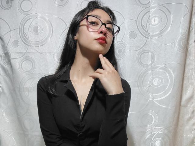 CamiVargas with black glasses, red lipstick, and black button-up shirt, touching her chin with her right hand, looking slightly up. She has long black hair and pale skin.