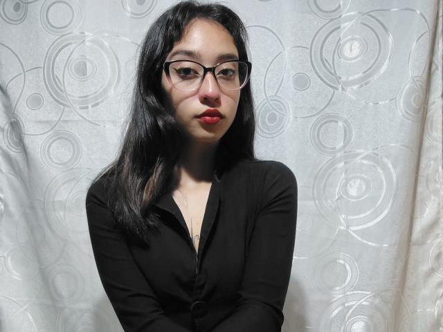 CamiVargas with long black hair, glasses, and red lipstick, wearing a black button-up shirt, standing against a white, circular-patterned curtain. Her ass is not visible.