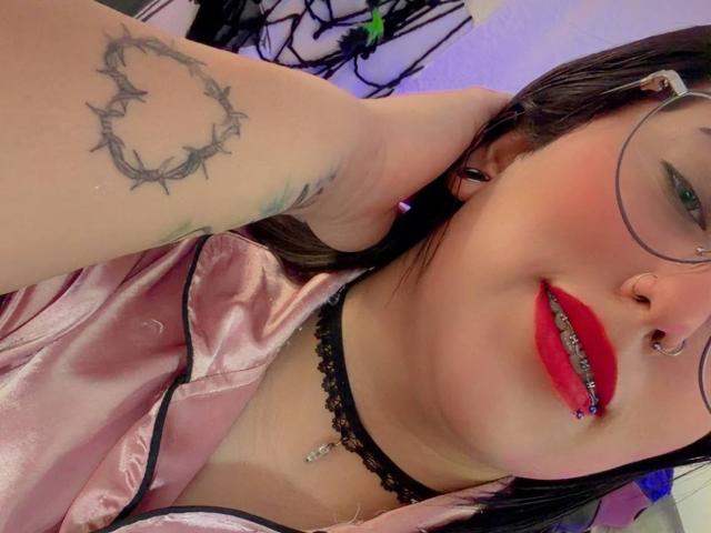 NatalieHarris with dark hair, red lipstick, nose ring, and glasses, wearing a pink satin top, black lace choker, and showing a thorn tattoo on her arm.