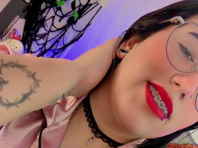 Natalie Harris, con lápiz labial rojo, collarín negro, gafas, piercing en la nariz y piercing en el tabique, mostrando sus dientes, tiene un tatuaje en el brazo y aretes negros, y lleva una camisa rosa..