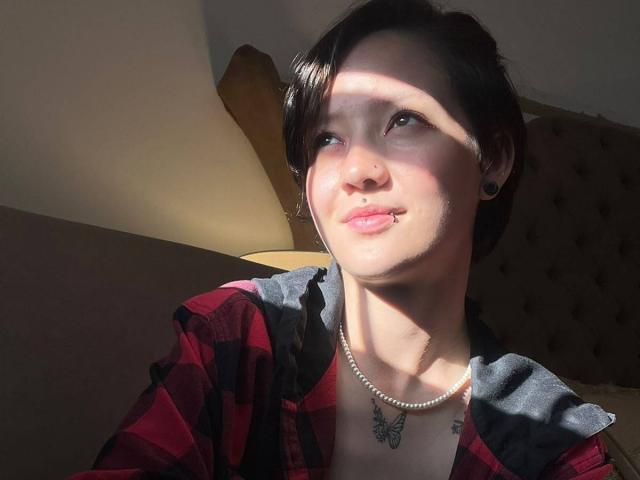 LissyLuxe has short black hair, a nose ring, black ear gauges, a pearl necklace, and a butterfly tattoo. She's wearing a red plaid shirt, smiling in sunlight.