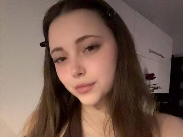 MaidaCain has pale skin, brown eyes, and long brown hair with a black hair clip, slight smile, and small nose, wearing a strap, looking slightly to the side.