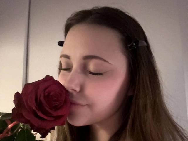 MaidaCain with eyes closed, gently kissing a deep red rose, holding it near her face. She has long brown hair, a black hair clip, and a soft smile.