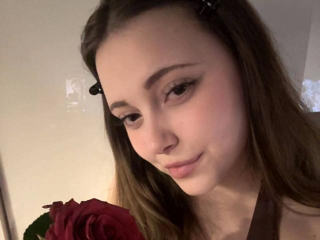 MaidaCain has a cute face with long brown hair, black hair clips, and a red rose in front, smiling softly with subtle makeup and a gentle, attractive look.