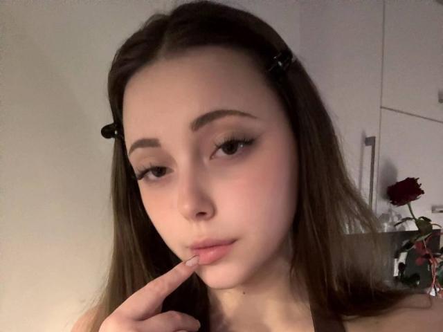 MaidaCain with light skin, brown hair in black hair clips, and makeup, biting her finger with a slight pout, showing off her small, pink, slightly parted lips.