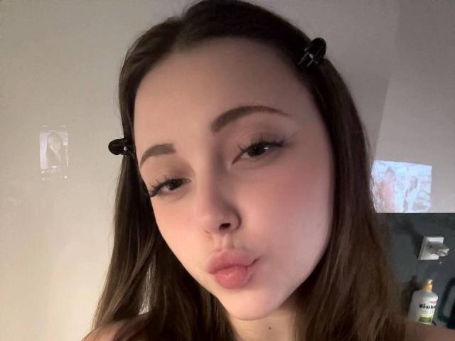 MaidaCain has light skin, brown hair in pigtails with black clips, and is making a pouty face with her lips slightly puckered. Her eyebrows are thick and she has long eyelashes.