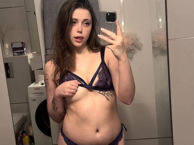 MaidaCain with long brown hair, wearing a dark purple lace bra and matching panties, showing her tits and holding a phone in a mirror selfie, has tattoos on her arm.