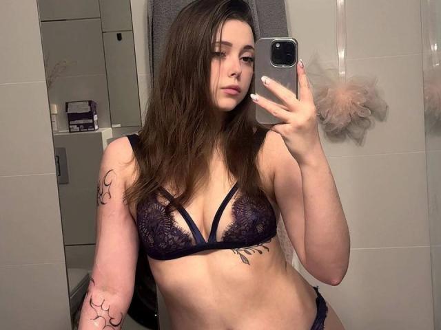 MaidaCain with brown hair, wearing a black lace bra and panties, holding a phone, showing tattoos on her arm and side, standing in a bathroom. Boobs, ass visible.
