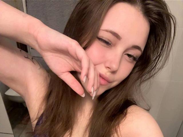 MaidaCain with brown hair, fair skin, and long nails, gently touching her cheek with a slight smile, showing her small tits and pink nipples in a bathroom selfie.
