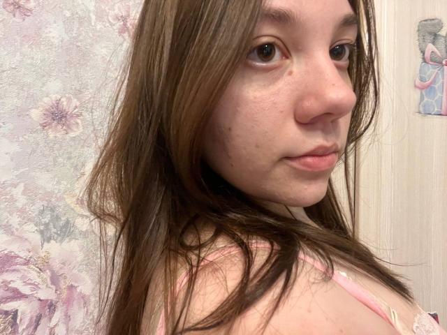 AureliaEmswiler has long brown hair, fair skin, and small freckles, wearing a pink lace top, looking over her shoulder with a slight pout. Her tits are partially visible.