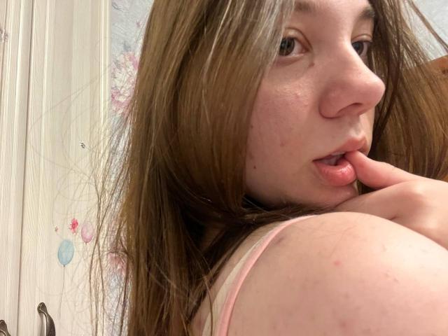 AureliaEmswiler with long brown hair, pink tank top, and slightly parted pink lips, touching her finger to her mouth, showing a small bruise on her shoulder.