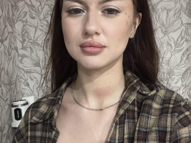 MarisaLipschutz has pale skin, brown eyes, and pink lips. She's wearing a plaid shirt with a chain necklace, and her brown hair is down. She's looking at the camera.