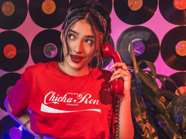 StefaValmont, portant une chemise rouge Coca-Cola, tenant un téléphone rouge, avec des seins de taille moyenne, des cheveux foncés attachés en un chignon désordonné, un anneau au nez et du rouge à lèvres rouge, se tient à côté d'une plante verte..