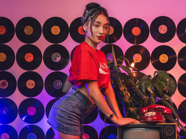 StefaValmont with dark hair in a bun, red t-shirt, and denim shorts, leaning on a TV, wearing a red phone bracelet, surrounded by vinyl records, in a pink-blue light.