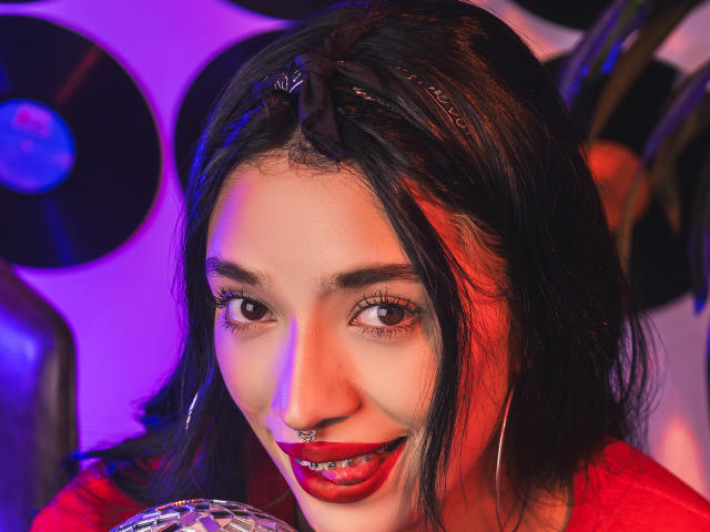 StefaValmont, avec ses cheveux noirs, son rouge à lèvres rouge, son anneau de nez et ses boucles d'oreilles, sourit à l'appareil photo. Des lumières violettes et bleues mettent en valeur son visage, et elle tient un objet scintillant..