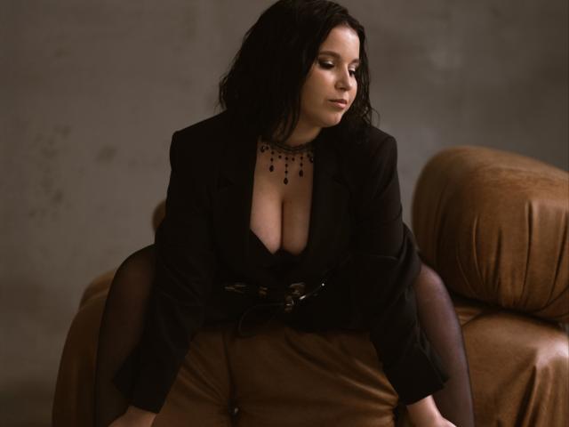 Simerria with black hair, wearing a black blazer and deep cleavage, sitting on a brown leather couch, legs apart, wearing sheer black stockings and a black necklace.