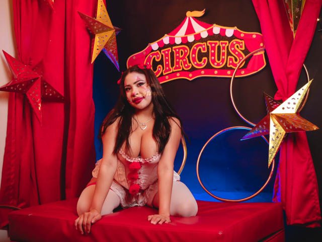 OliviGrace, with big tits and a cute face, kneels on a red cushion, wearing pink lingerie and white stockings, in a circus-themed setup with red curtains and star decorations.