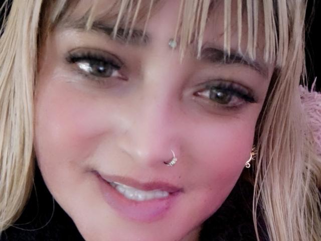 AlessadraJoness has blonde hair, green eyes, pinkish skin, a nose ring, and a small dot on her forehead. She's smiling with pink lips. Her bangs are straight.
