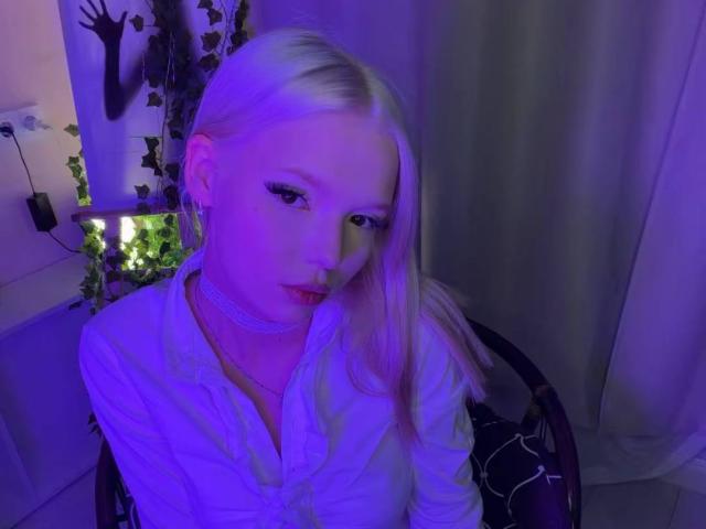 Willodean Almonte, con cabello rubio platino, vestida con una camisa blanca, rostro iluminado en color púrpura y un collar, mirando hacia abajo, sentada en una silla, con una planta verde en el fondo..