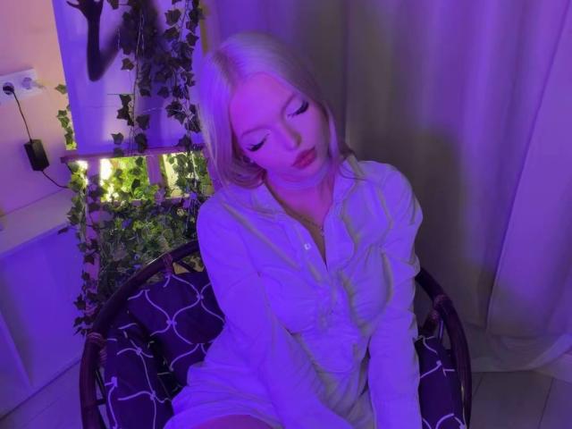 Willodean Almonte mit blonden Haaren, im violetten Licht, sitzend auf einem Stuhl, trägt ein weißes Hemd mit Knopfleiste, blickt nach unten, Hände auf den Knien, violette Schatten auf dem Gesicht, Pflanze im Hintergrund..