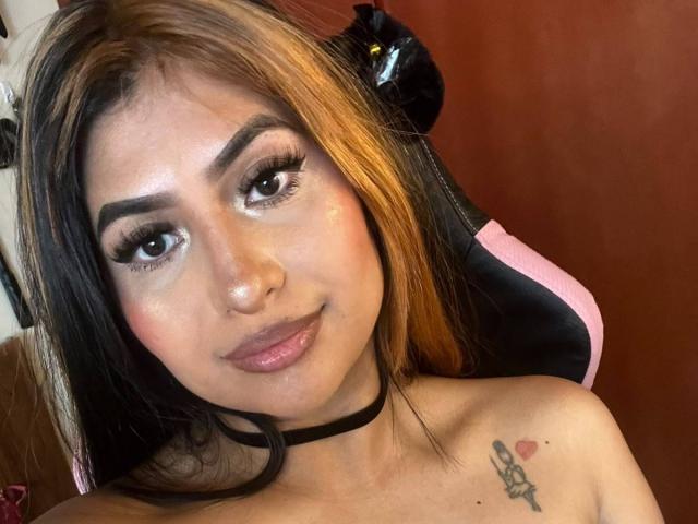 ZarayWhitMoreLove with shoulder tattoos, black choker, and pink and black hair, sitting in a gaming chair, has a cute smile and eyeliner, showing her tits.