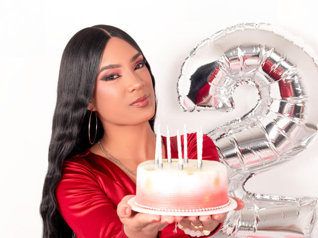 NatLewis with long black hair, red satin shirt, holding a white cake with candles, and a silver number two balloon, smiling slightly. Boobs visible under shirt, hoop earrings.