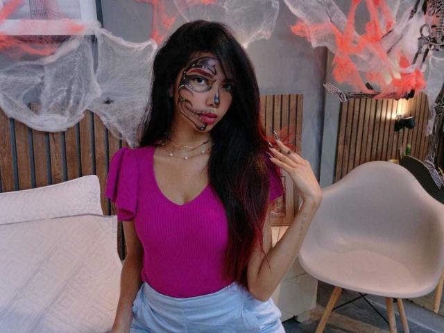 LunaRoja69 with long black hair, pink top, white pants, facial paint, holding hair, pink chair, white pillow, wooden wall, and spider webs. Her tits are small and her ass is hidden.