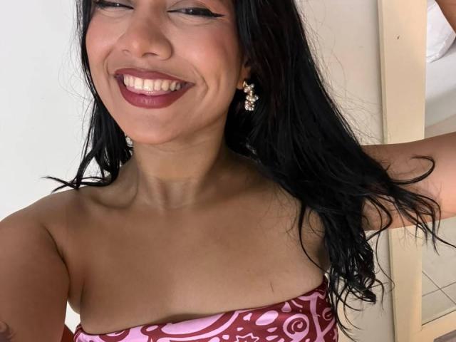 AlanaLuxe, avec ses cheveux foncés, souriante, portant un haut sans bretelles à motifs bordeaux et rose, dévoilant sa poitrine, avec des boucles d'oreilles en diamants et du rouge à lèvres rouge foncé..