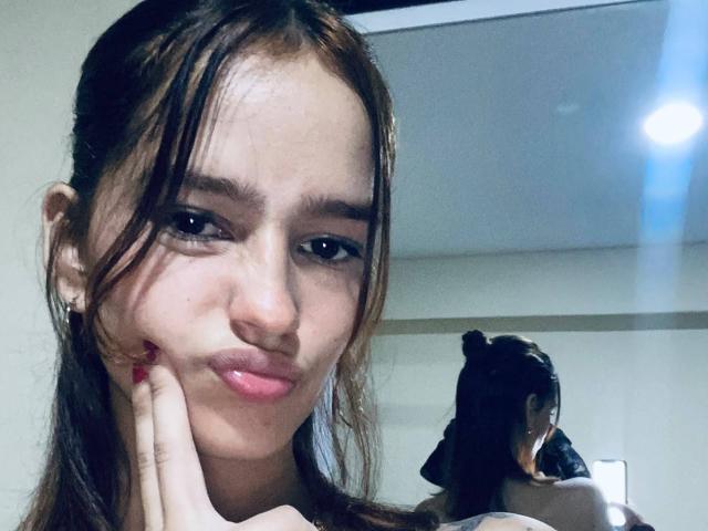 MelodyMansson has pouty pink lips, brown eyes, and long brown hair, with her hand on her cheek, looking at a mirror reflection showing her back and top bun.