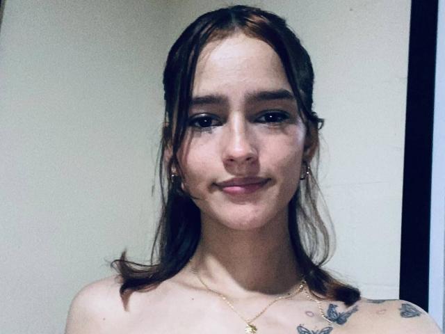 MelodyMansson has a cute face, brown hair, small tits, tattoos on her right shoulder, and a subtle smile. She's wearing a gold necklace and earrings.