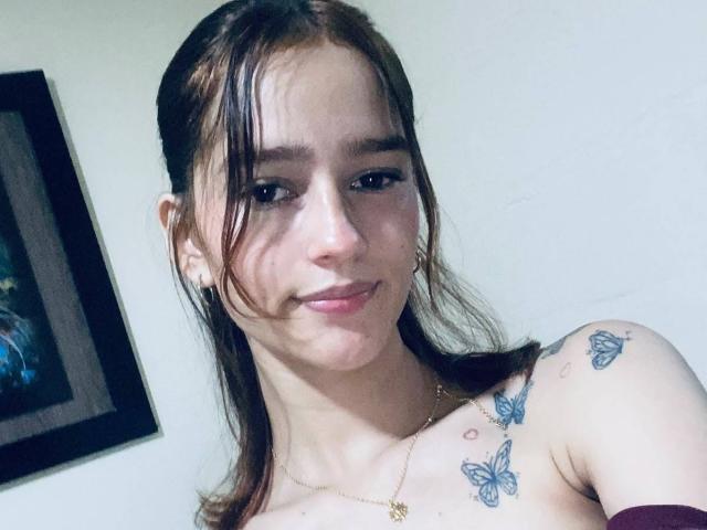 MelodyMansson with brown hair, light skin, and small tits, has butterfly tattoos on her shoulder, wearing a gold necklace, smiling slightly, in a selfie.