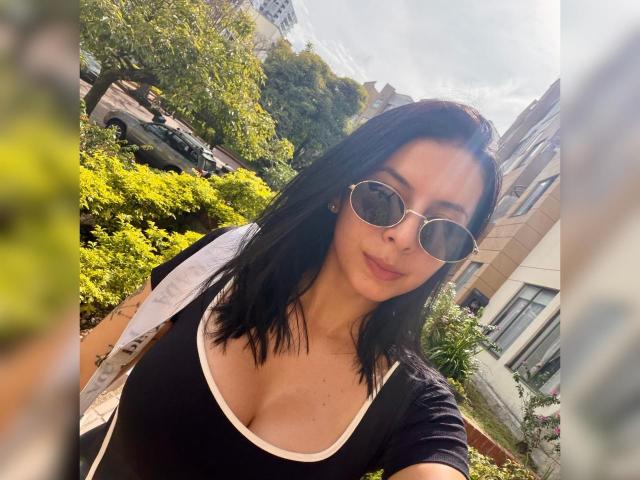 Isabella, con i capelli neri, indossa occhiali da sole rotondi e una maglia nera con bordi bianchi, che mette in mostra il seno, ed è in piedi all'aperto, in una giornata di sole, tenendo in mano una borsa bianca..